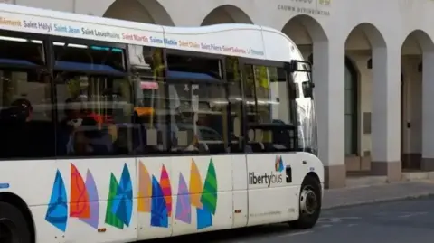 BBC A LibertyBus driving down the road. It is a white bus with multicoloured shapes along the bottom. It has large black windows.