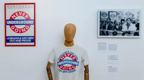Mannequin wearing a white T-shirt reading “Never Going Underground: Lesbians & Gay Men Out and Proud”, displayed beside campaign posters and photographs.