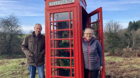 Kathy Cook and Roger Deeks standing beside the restored phone box. The surrounding area has been cleared of brambles and bracken, and the phone box has been painted a bright red, with the Telephone signs newly installed. Kathy is wearing a padded purple coat and glasses and is leaning out of the open door, smiling at the camera. Roger is standing on the left, wearing blue jeans and a brown jacket with his hands in his pockets. It is a sunny winter day and the trees are bare.