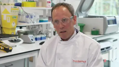 Darius Hughes has short grey hair. He is sat in a lab wearing safety goggles and a white lab coat with a Moderna logo on it.