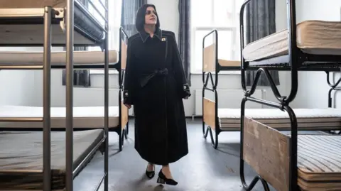 PA Media Shabana Mahmood walks through a dormitory room which contains bunk beds to her left and right. She is wearing a black coat and black shoes.