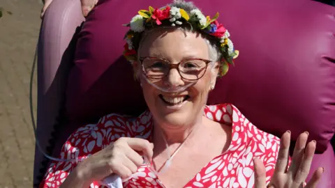 St Elizabeth Hospice A woman with glasses and a colourful dress on sitting on a large pink chair outside. She has flowers in her hair and tubes coming from her nose.