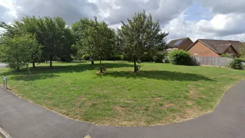 Google Street View of the land in Church Walk. It is a grassed open space with several trees dotted around, screening off houses in the background.