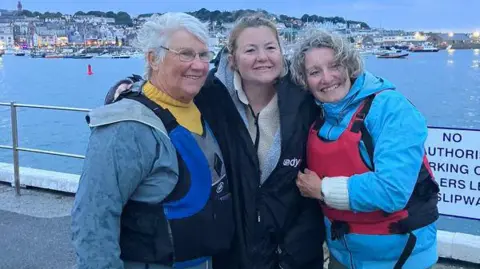 BBC Rosie Williams is being hugged by two women at dusk in the harbour.