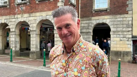 Matt Whittaker is wearing a multi-coloured paisley-patterned shirt and standing outside Torrington Town Hall