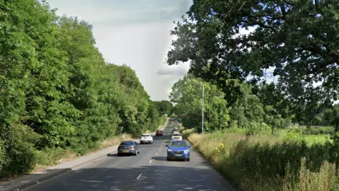 Google streetview image of an A-road with trees and field either side.
