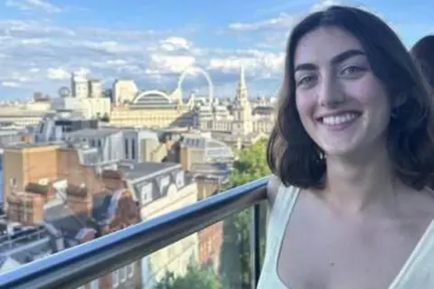 Eleanor Wallis Eleanor Wallis is wearing a white top and is smiling at the camera. There is a view of London on a partly cloudy day behind her.