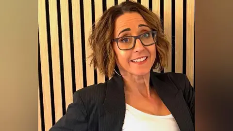 Natalie Queiroz A woman with short, light-brown hair and dark blue glasses. She is wearing a black pinstriped suit jacket with a white top and is standing in front of a wood panelled wall.