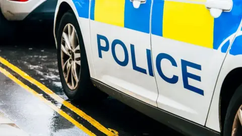 The side of a police car, with "police" emblazoned on it in blue on a white background, parked on the side of a wet road.