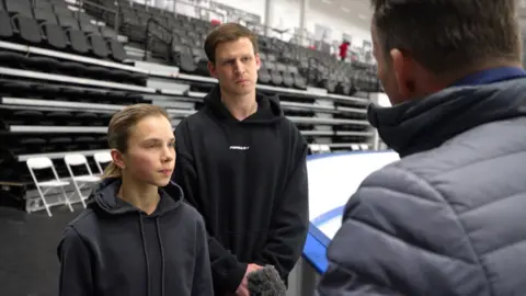 In black sweatshirts, women and men stand in front of empty ice rink seats next to the microphone, behind man's head