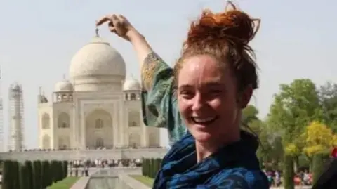 Danielle McLaughlin stands in front of the Taj Mahal - it can be seen in the background and she is holing her right hand up pretending to rest her finger on the top of the Taj Mahal's dome. He red-brown hair is tied up in a bun on top of her head.