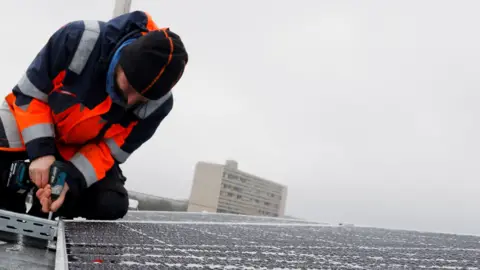 Reuters A stock image of solar panels being installed on a roof. An engineer wears a hi vis jacket and uses a drill. 