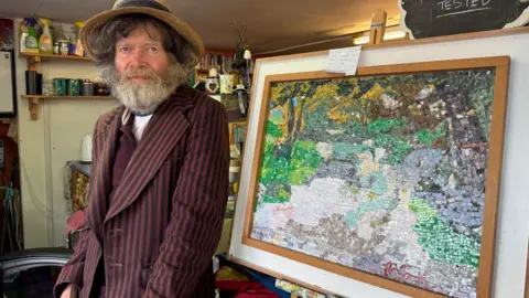 Artist, Paul Hoare, is 69 and closed his art Gallery in Cardigan last week because of rising costs he is pictured next to one of his mosaics and wears a straw boater and striped blazer and has a beard.