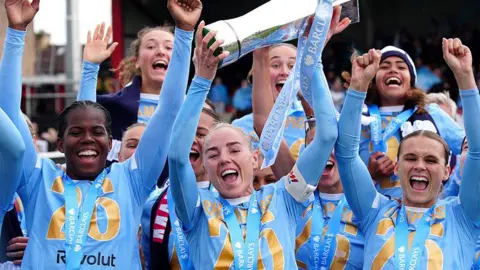 PA Media Manchester City's Alex Greenwood lifts the trophy with team-mates following winning the Women's Super League at the Chigwell Construction Stadium, Dagenham. The players are all cheering with their hands in the air.