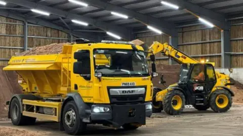 Two yellow gritter vans filling up their vehicles with grit from a warehouse.