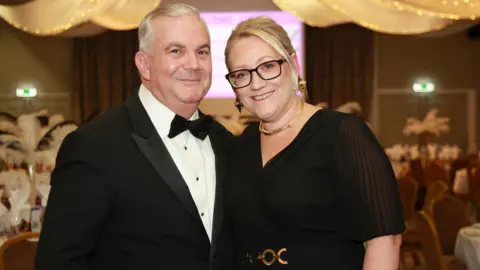 David and Jo Ward pictured in formal attire. David, who is on the left, is wearing a black tuxedo with a black bowtie. Jo, on the right, is wearing a black dress with a gold belt feature. 