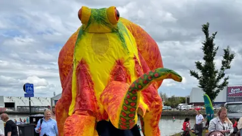 North East Lincolnshire Council A person wearing a large green, orange and yellow sea creature costume. People are walking the streets behind and the sky is overcast. 