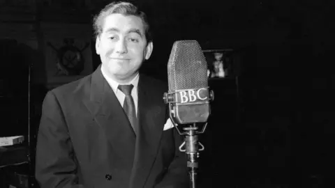 A black and white image of comedian Tony Hancock in a suit, smiling, near a microphone that says "BBC" on it. 