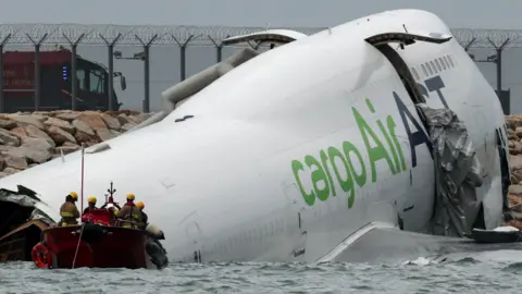 Reuters Part of a white coloured plane's fuselage lies in the water by a runway as a vessel with rescue officials sail towards the wreckage. The emergency doors are open and you can see grey emergency chutes protruding from the doorways.