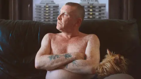 Dumbworld Productions  Joe McNally in The Flats pictured topless on sofa with dog and New Lodge flats visible through window behind him