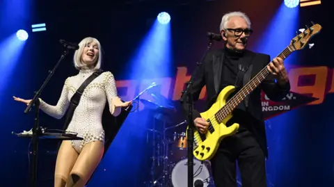 PA Media Singer Sophie Grey (left) and Trevor Horn performing. Grey has a blonde bob and is wearing a a pearl-coloured outfit. Horn has short grey hair, brushed back from his face, and is wearing a black suit and top as he plays bass guitar. The stage is illuminated by blue spotlights.
