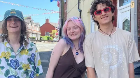 Jonny Humphries/BBC Three teengers, two men and one woman, smile at the camera on a street with flags and bunting in the background