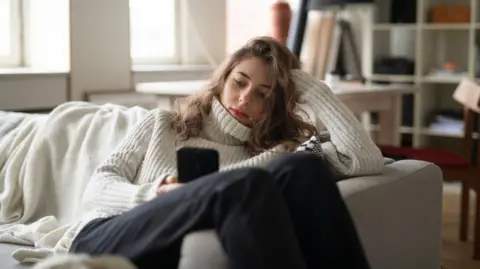 Getty Images A young woman wearing a high-necked white jumper, sitting on a sofa with her legs over the side, looks at a phone