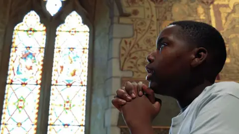 Wycombe Youth Action / Love Music Hate Violence A close-up side profile of a person seated inside a church, with both hands clasped beneath the chin. The person is wearing a white T-shirt and is positioned in the right foreground of the image. Behind, two tall stained-glass windows fill the left side of the frame, casting bright natural light into the space. Decorative painted walls, stone arches and ornate religious artwork surround the windows, highlighting the historic interior. The composition contrasts the sharply focused foreground figure with the softly illuminated church background, drawing attention to the architectural details and the reflective pose.