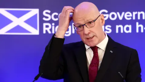 PA Media John Swinney, first minister of Scotland, scratching his head at a press conference. He is weating a dark suit, white shirt and burgundy swirly tie. He is wearing an enamel saltire badgs on his left lapel.