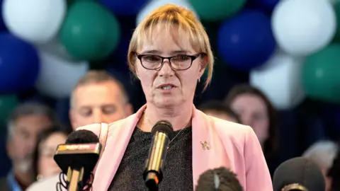 Sandie Peggie in pink jacket and grey blouse, wearing glasses, in front of a mic. There are people behind her.