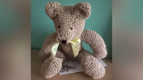 A beige string teddy bear with a yellow bow against a green wall. A yellow ribbon is around its neck.