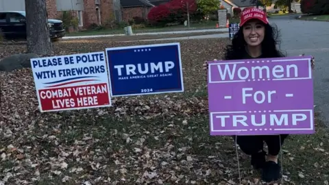 Amanda Sue Mathis Amanda Sue Mathis mengenakan topi Make America Great Again berwarna merah sambil berjongkok di balik tanda Women for Trump berwarna ungu di halaman rumahnya yang dipenuhi dedaunan musim gugur. Tanda lainnya termasuk tanda kampanye Trump angkatan laut dan tanda yang bertuliskan: "Harap bersikap sopan dengan kembang api. Veteran tempur tinggal di sini."