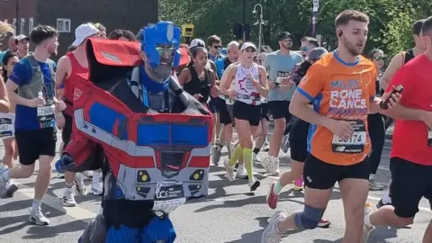 Andrea Hudson Matt Batchelor runs in a crowded road race, wearing a bright red and blue Optimus Prime costume, surrounded by other runners in sports clothes on a sunny, tree-lined street.