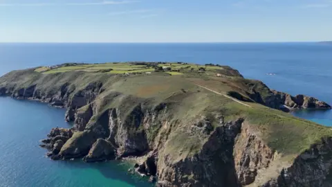 A birds eye view of Sark. There is a dirt path and lots of green fields. You can see the edges of the Island and surrounding sea