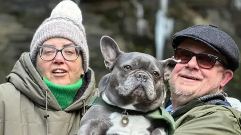 Maria Elliott A man and woman hold a grey French Bulldog up between them. On the left, the woman wears a grey woolly hat, glasses, a green turtleneck and a light green waterproof. The man wears a black flat cap, dark sunglasses and a green waterproof. He has a short grey beard.