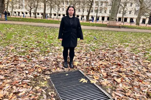 Snezana Grigorjeva is seen in a black coat stands on autumn leaves near a ground-level light grille in a city park. Behind her are leafless trees, a historic building, and a bench.