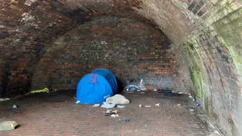 A blue tent pitched inside a railway arch with some rubbish strewn across the floor.