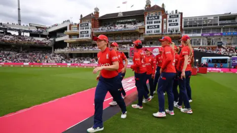 Getty Images England women cricket players in red jerseys and caps walk across the field in front of a packed stadium, with the historic pavilion building visible in the background.