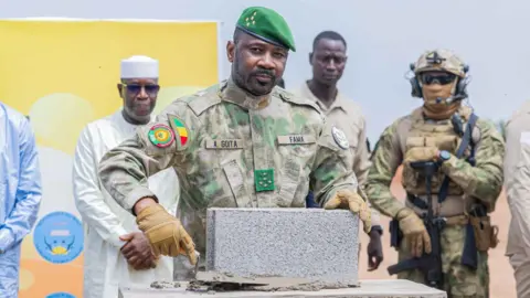 Gen Assimi Goïta, in military uniform, is standing in front of a brick at the construction site of the refinery in Senou, as armed soldiers in uniform and men in civilian clothes look on