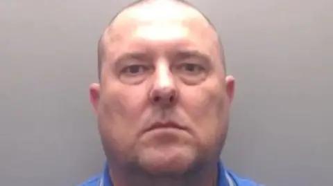Durham Constabulary A mugshot of a bald Jason Howe who is in his 50s with tattoos on his arms wearing a blue Boss branded T-shirt.