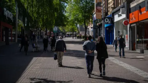 People are seen walking up and down the high street with shops on either side. The street has plenty of greenery and is well-kept.