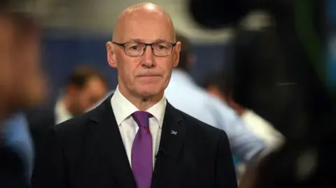 Getty Images John Swinney looking serious in a dark suit and white shirt with purple tie. People can be seen talking in the background.