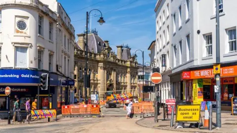 Getty Images An urban renewal project takes place in Wolverhampton in 2022. A road is closed for construction work and people are walking around the city. There are shops on both sides of the road, and there is a sign saying "businesses open as usual".