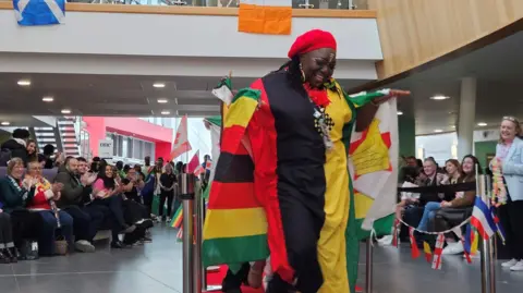 Eastern Education Group A woman walks down a cat walk in a multi-coloured outfit. She wears a red hat and is smiling. Seated spectators on either side are smiling and clapping