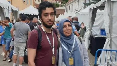 A man and a woman stand together at an outdoor market with white tents lining the street. Behind them are lots of people, tents and green hills under a cloudy sky.