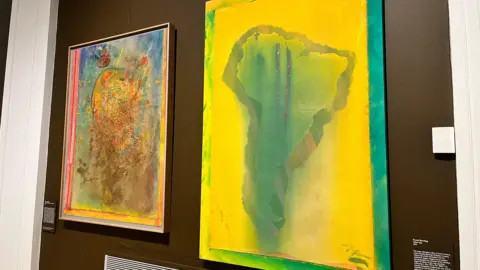 Emma Baugh/BBC Two paintings are side by side on the wall. The one on the left is smaller and is a contemporary piece which is made of several colours, including blue, yellow and brown. The one on the right is larger and is an outline of Africa.
