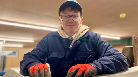 A teenager wearing glasses,  a cap, blue overalls and safety gloves leans over a bench,  facing the camera. 