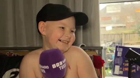 BBC Riley is holding a purple Radio Tees microphone and is smiling broadly at the camera. The nine-year-old is wearing a black cap.