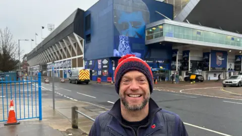 Luke Deal/BBC Tom Wright wearing a blue, red and white woolly hat and blue coat, he is stood in front of Portman Road stadium.
