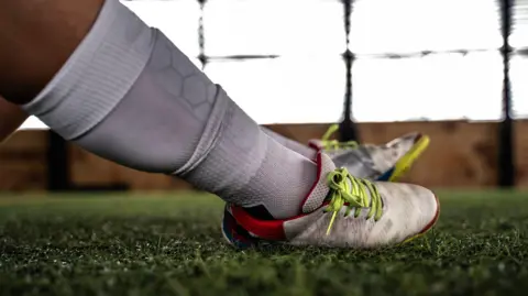 A close-up of someone's legs with white football socks and boots. They are sitting on a green artificial pitch.